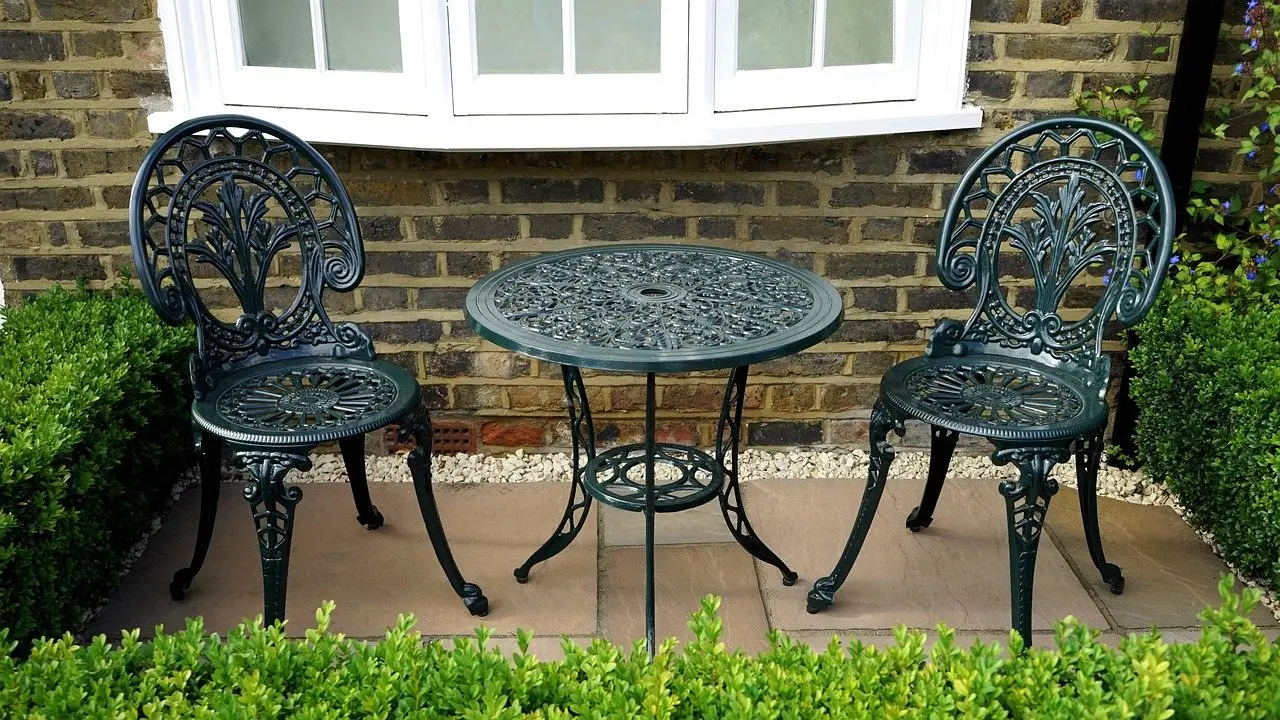 An ornate cast metal bistro table and two chairs on a paved patio against a brick wall
