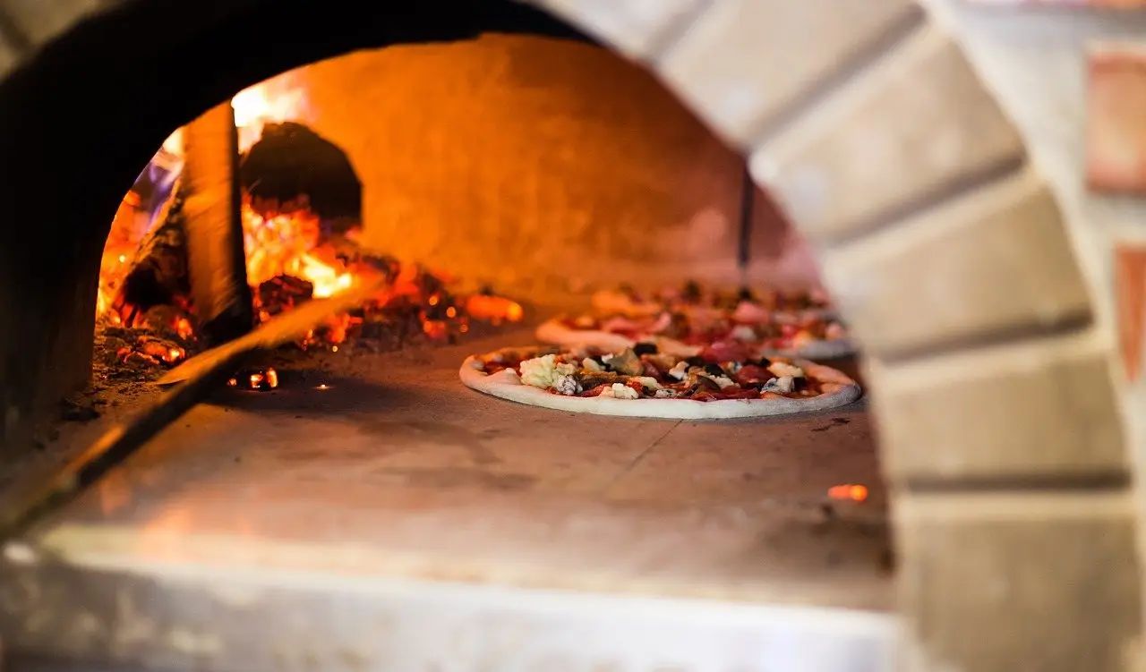 A pizza cooking on the stone floor of a brick wood-fired oven beside burning logs