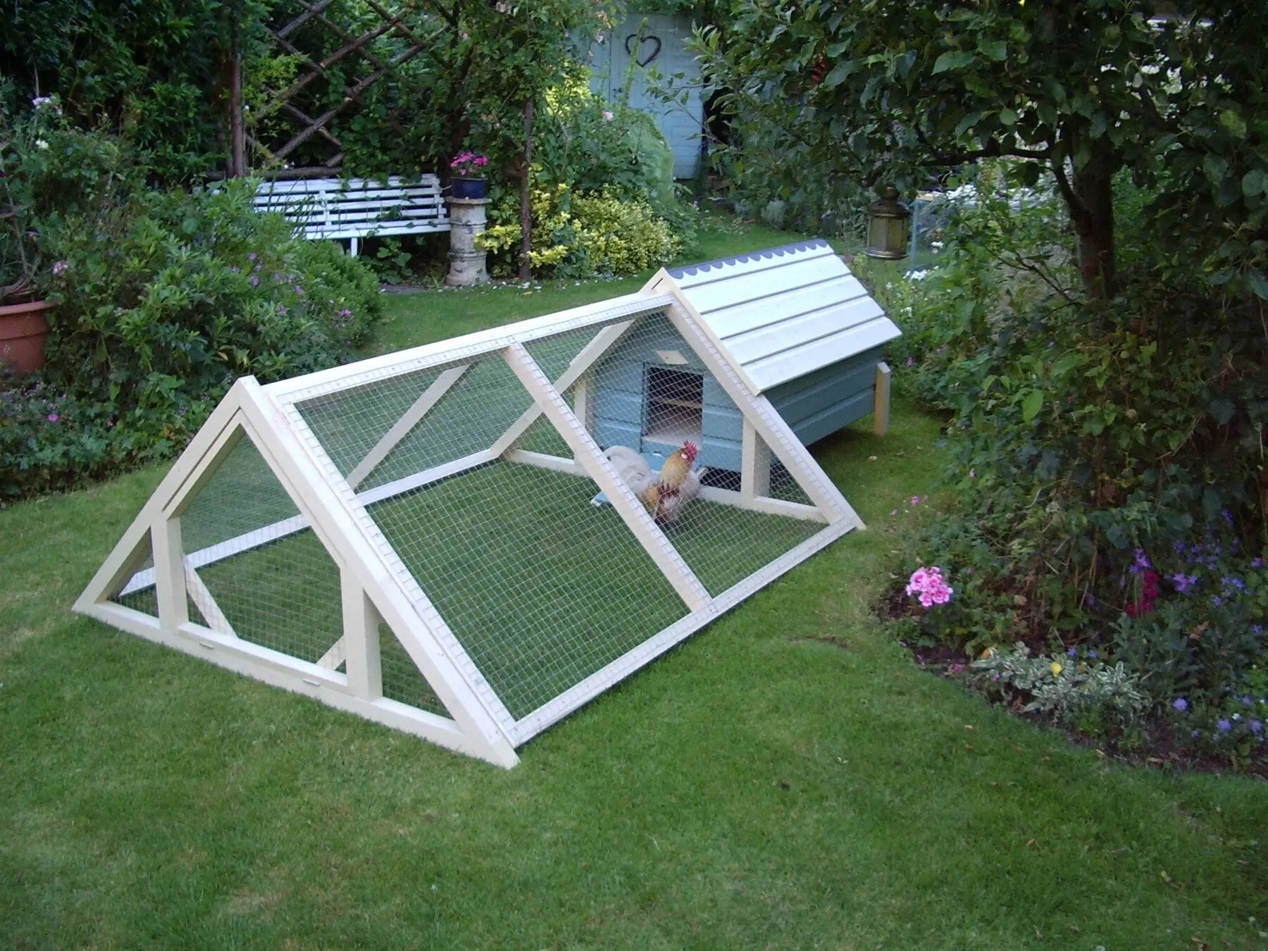 A small garden coop with a fully mesh-covered A-frame run attached, hens sheltering inside