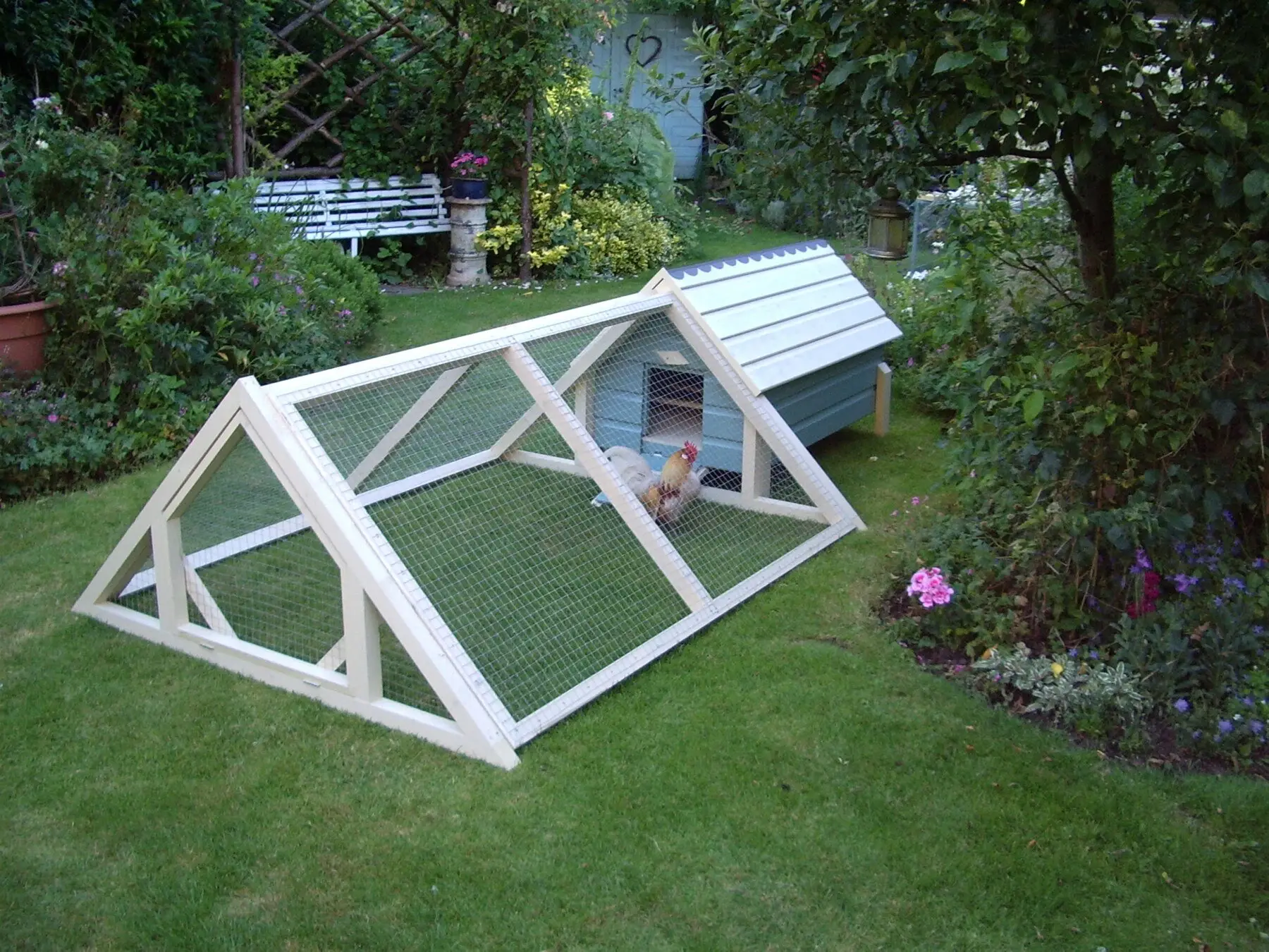 A small garden coop with a fully mesh-covered A-frame run attached, hens sheltering inside