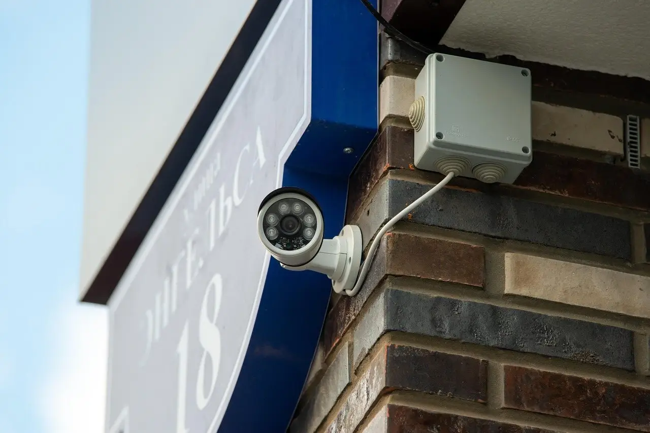 A bullet security camera mounted on a brick wall beneath an eave, cable running to a junction box