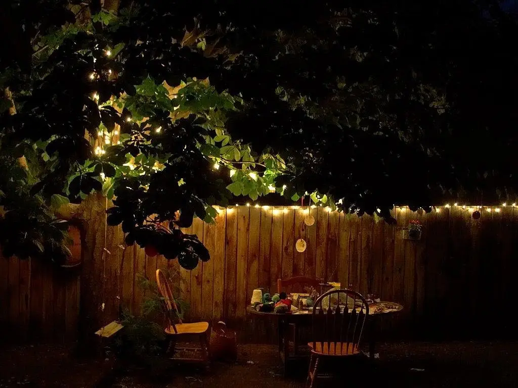 Warm string lights strung through a tree and along a fence above a lit garden table at night