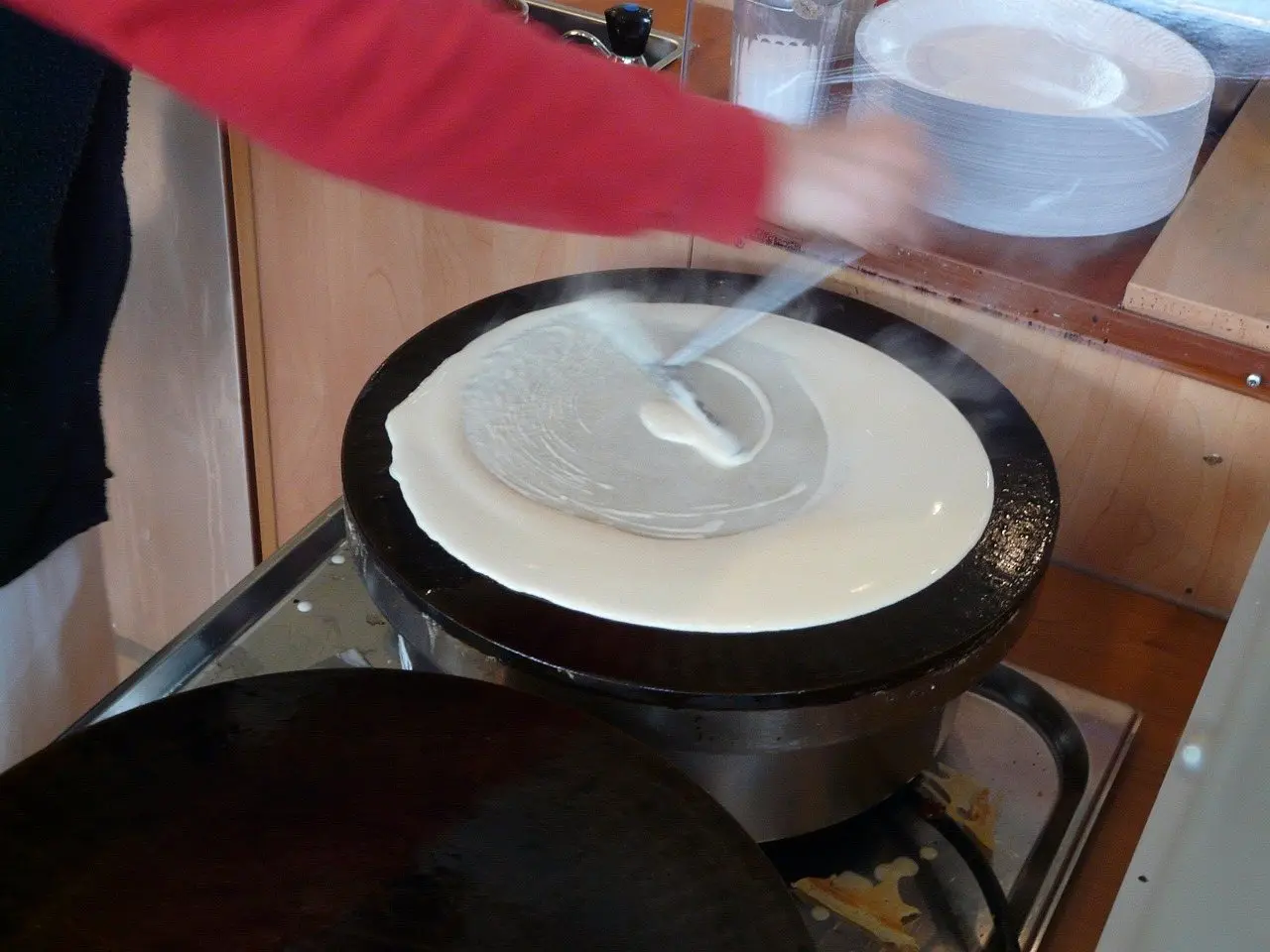 Batter being spread across a hot round griddle plate with a wooden spreader