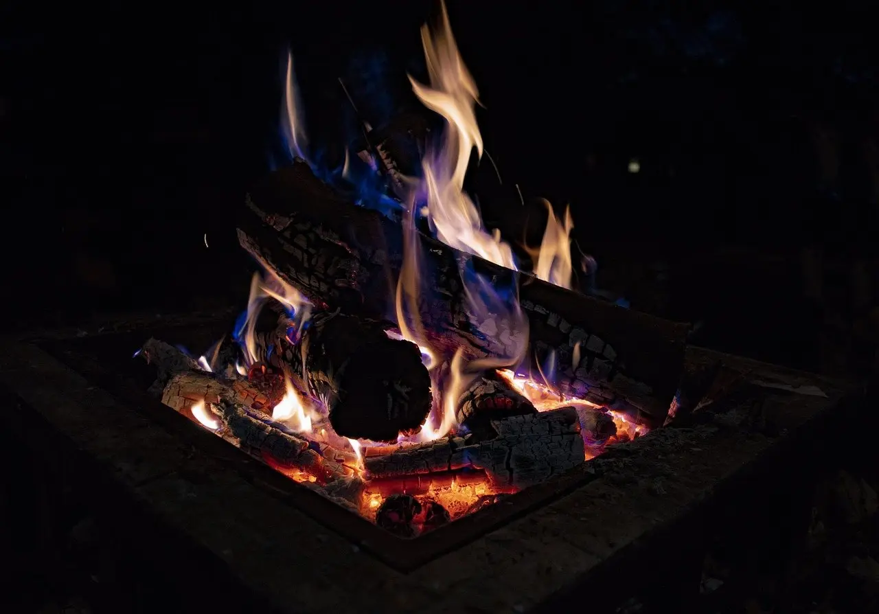 Logs burning with blue and orange flames in a square metal fire pit after dark