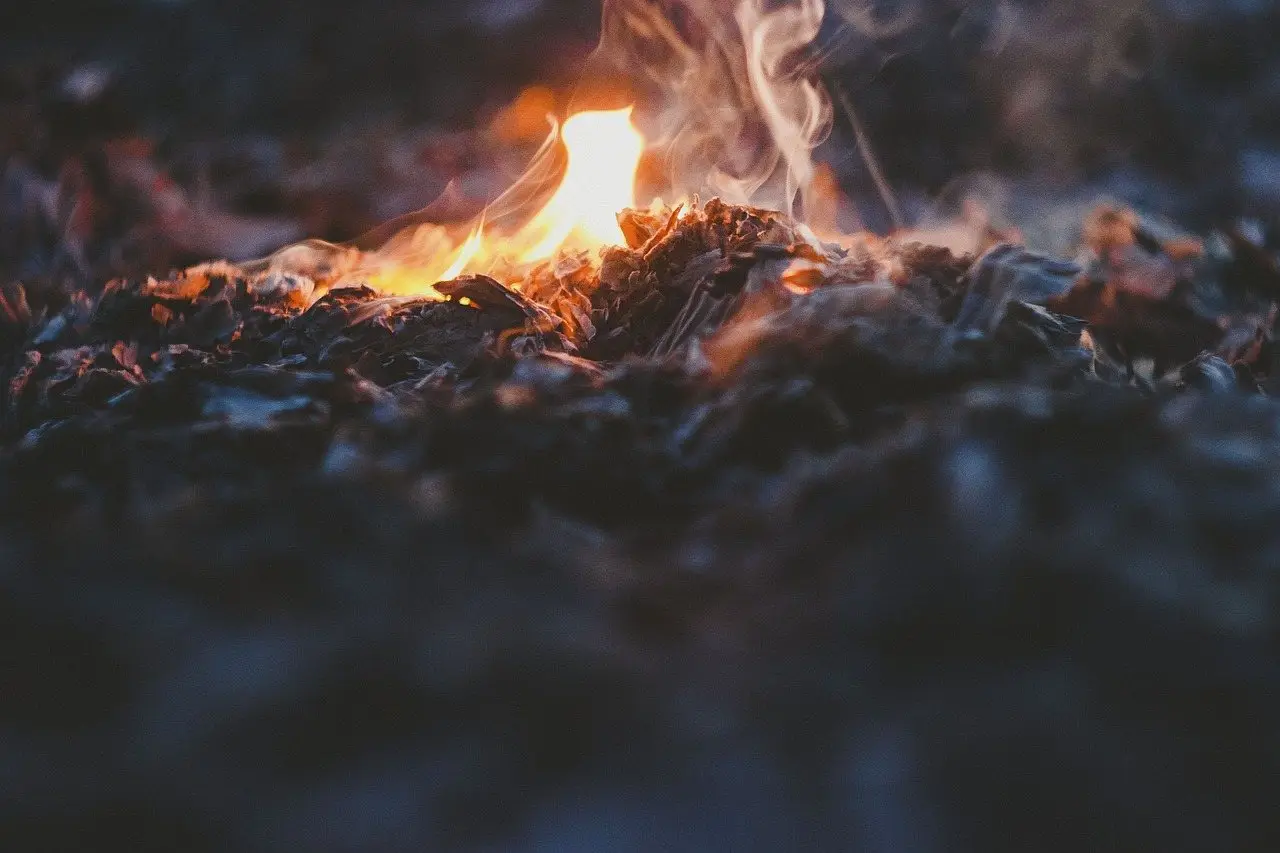 Pale smoke drifting from the glowing embers of an outdoor fire at dusk