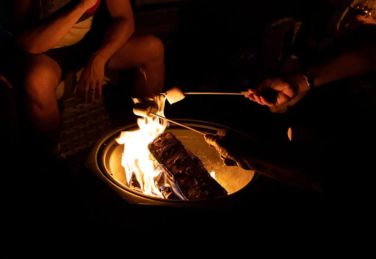 People seated close around a lit fire pit at night, holding roasting sticks