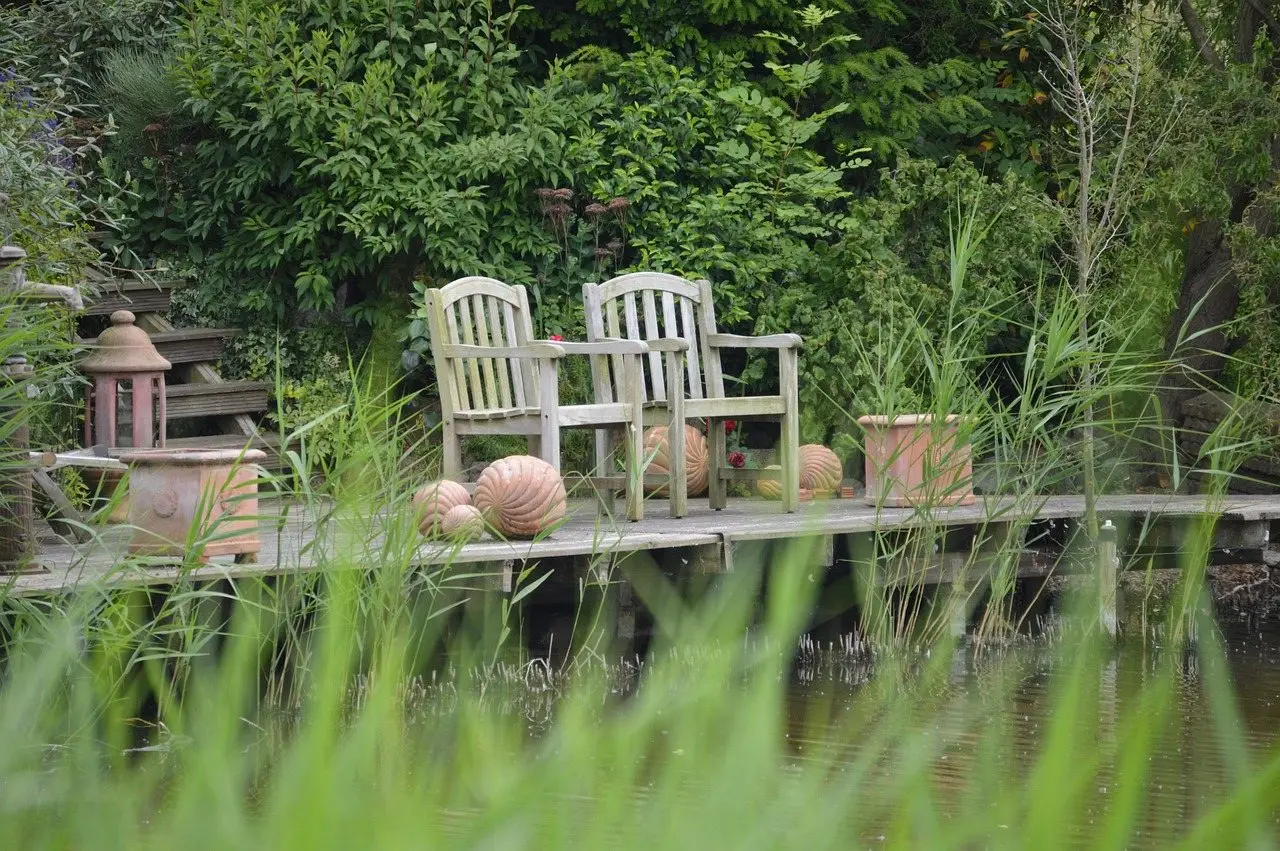 Two wooden chairs on a timber deck beside a pond, surrounded by planting