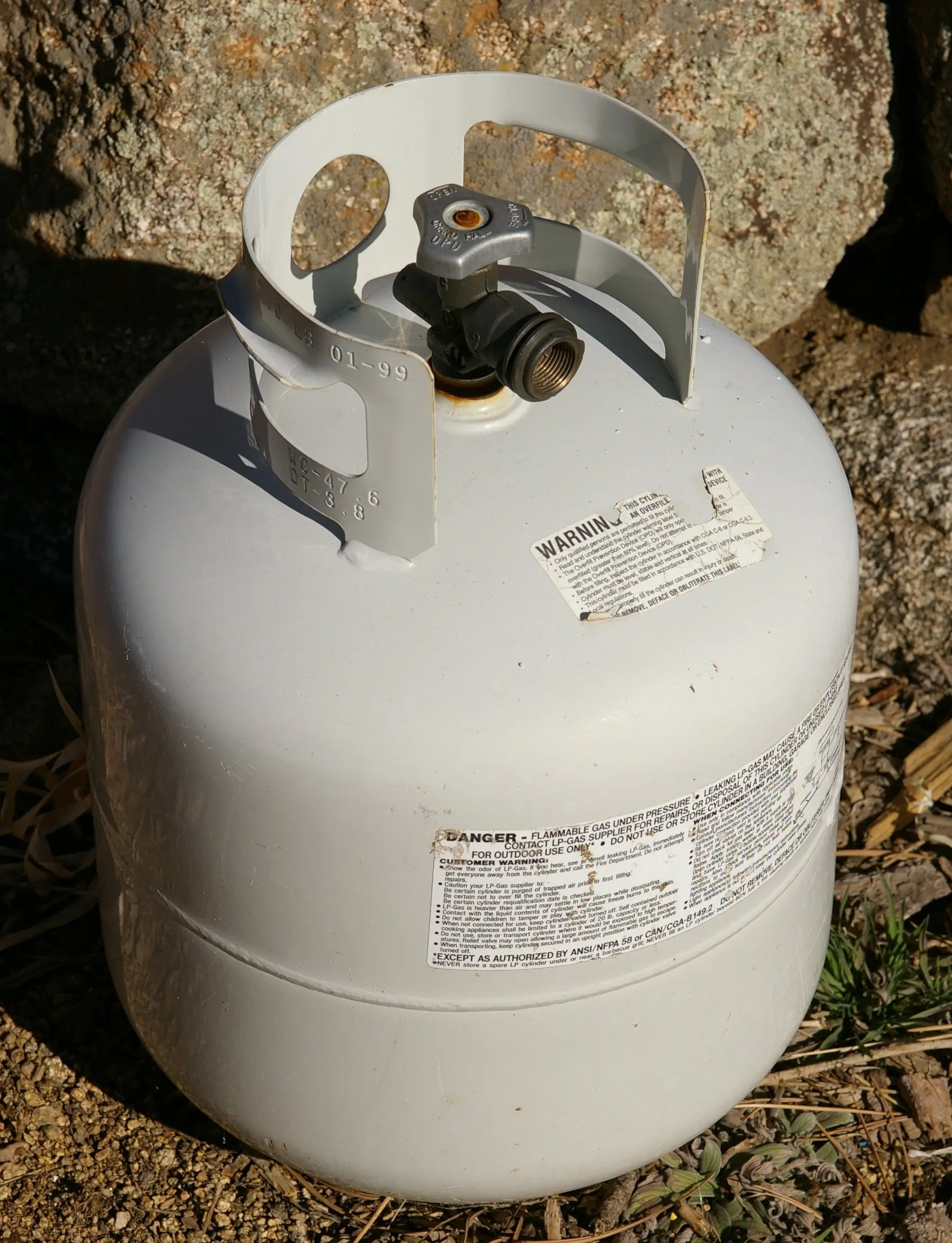 A standard 20 lb propane cylinder with its collar handle and service valve, standing on gravel