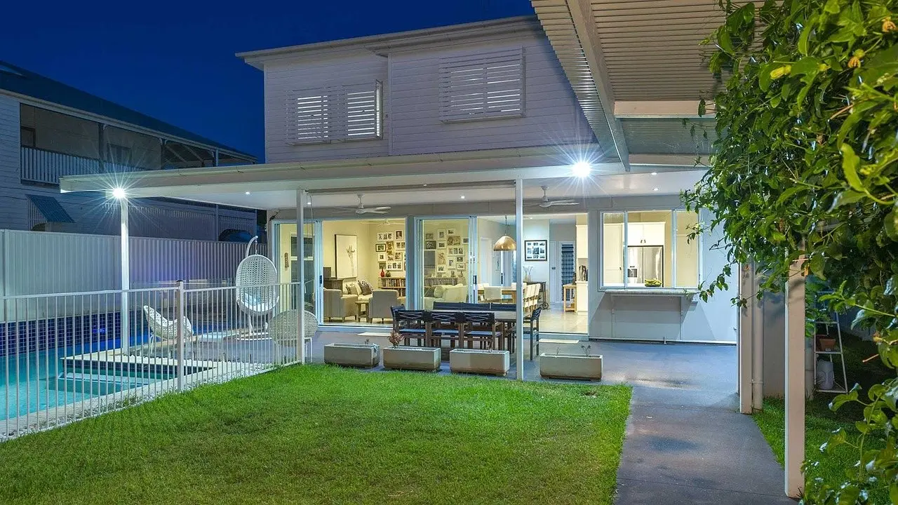The rear of a house at dusk with a covered terrace, lit interior, lawn and pool visible
