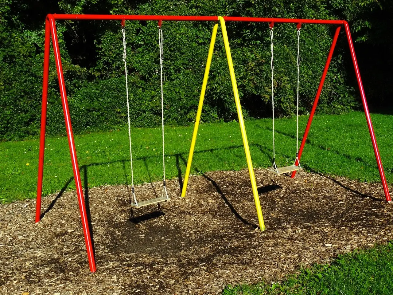 A red and yellow metal A-frame swing set with two seats, standing on a bed of bark chip