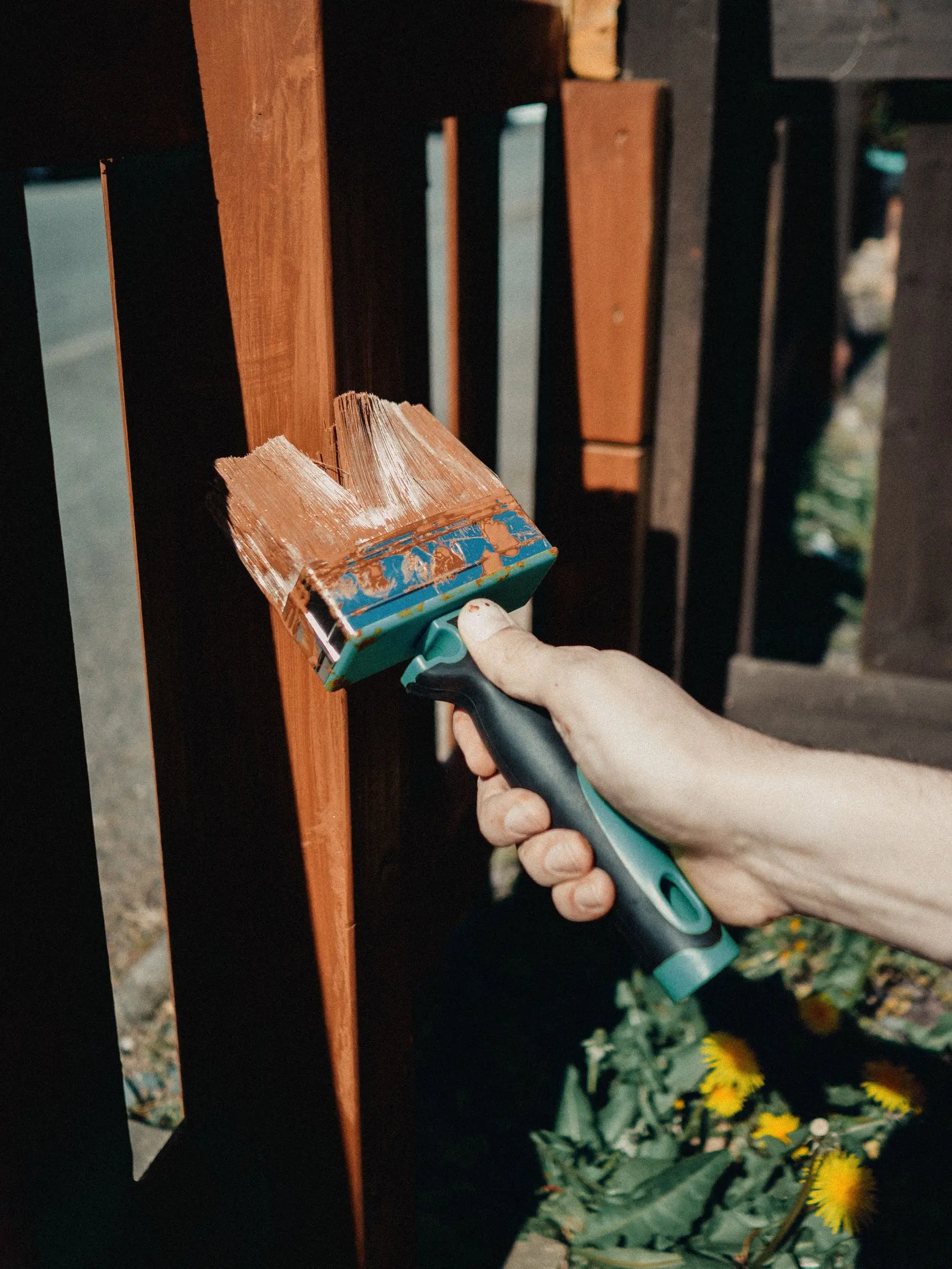 A hand brushing wood stain onto bare timber boards, with half of one board still unstained