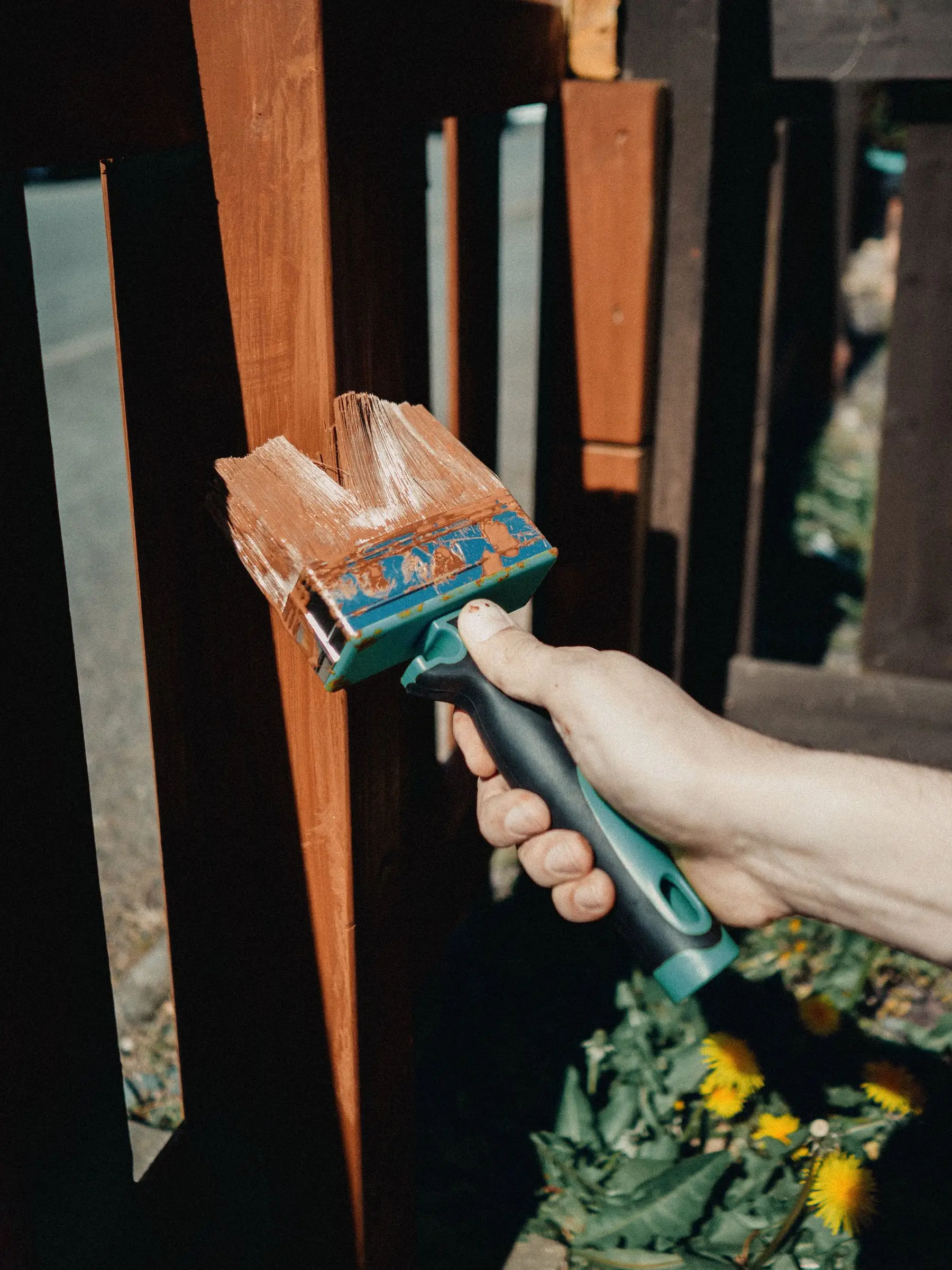 A hand brushing wood stain onto bare timber boards, with half of one board still unstained