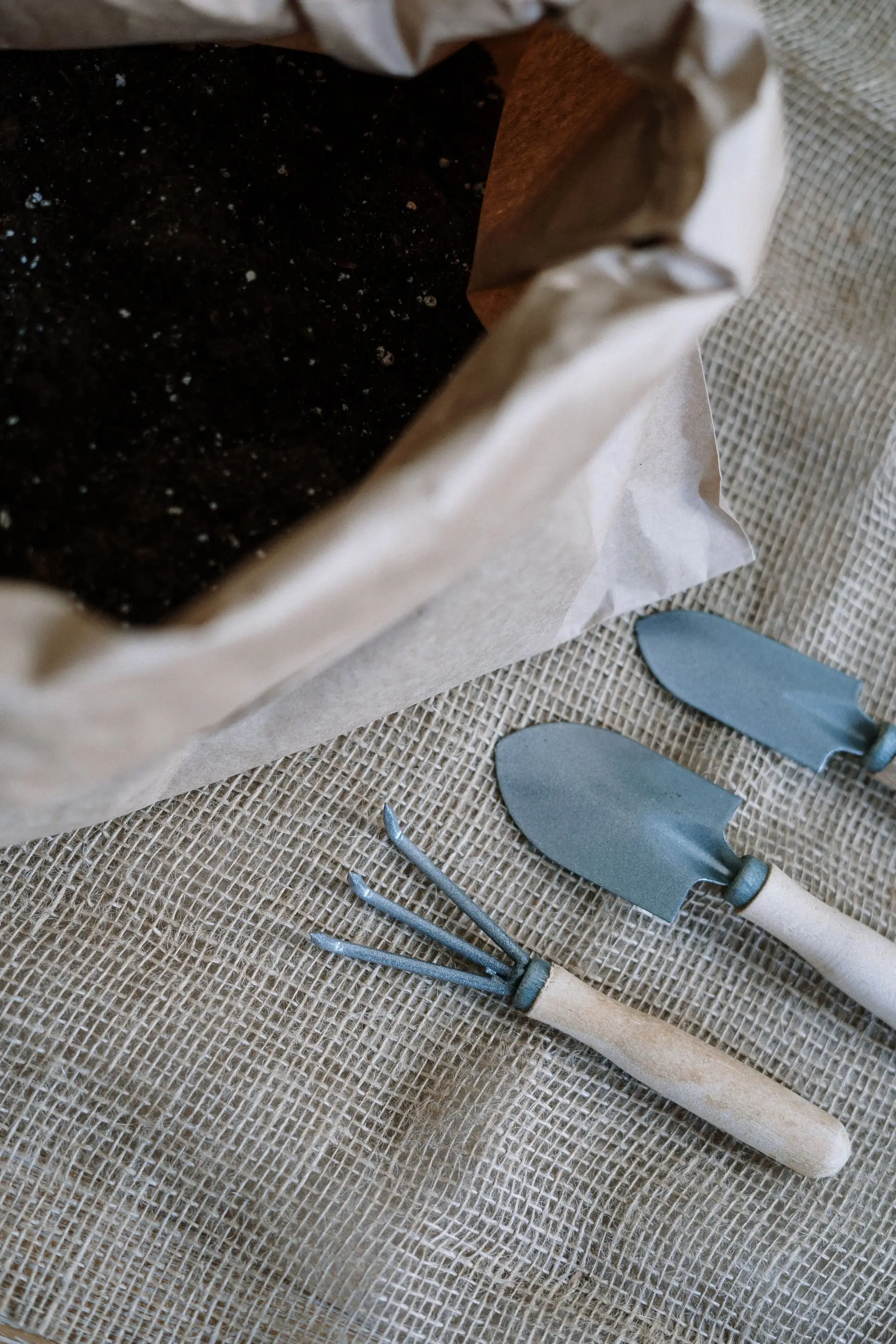 Hand trowels and a hand fork with wooden handles laid on burlap beside an open bag of compost
