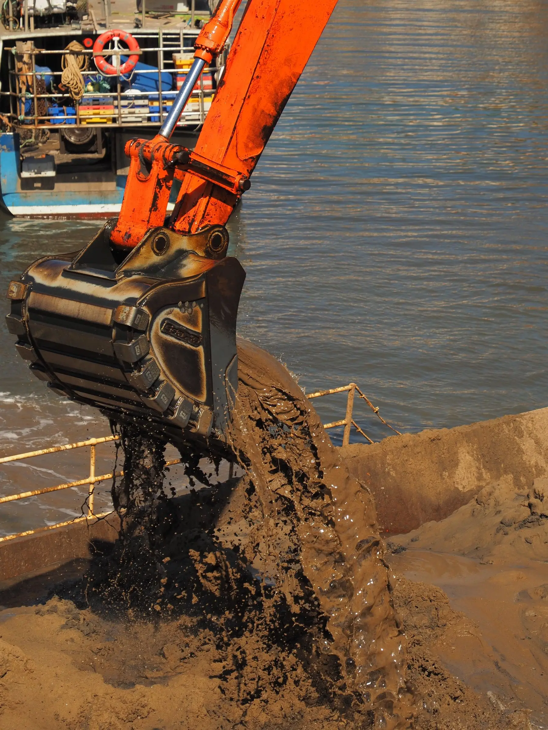 An excavator bucket tipping wet grey silt onto a bank during harbour dredging