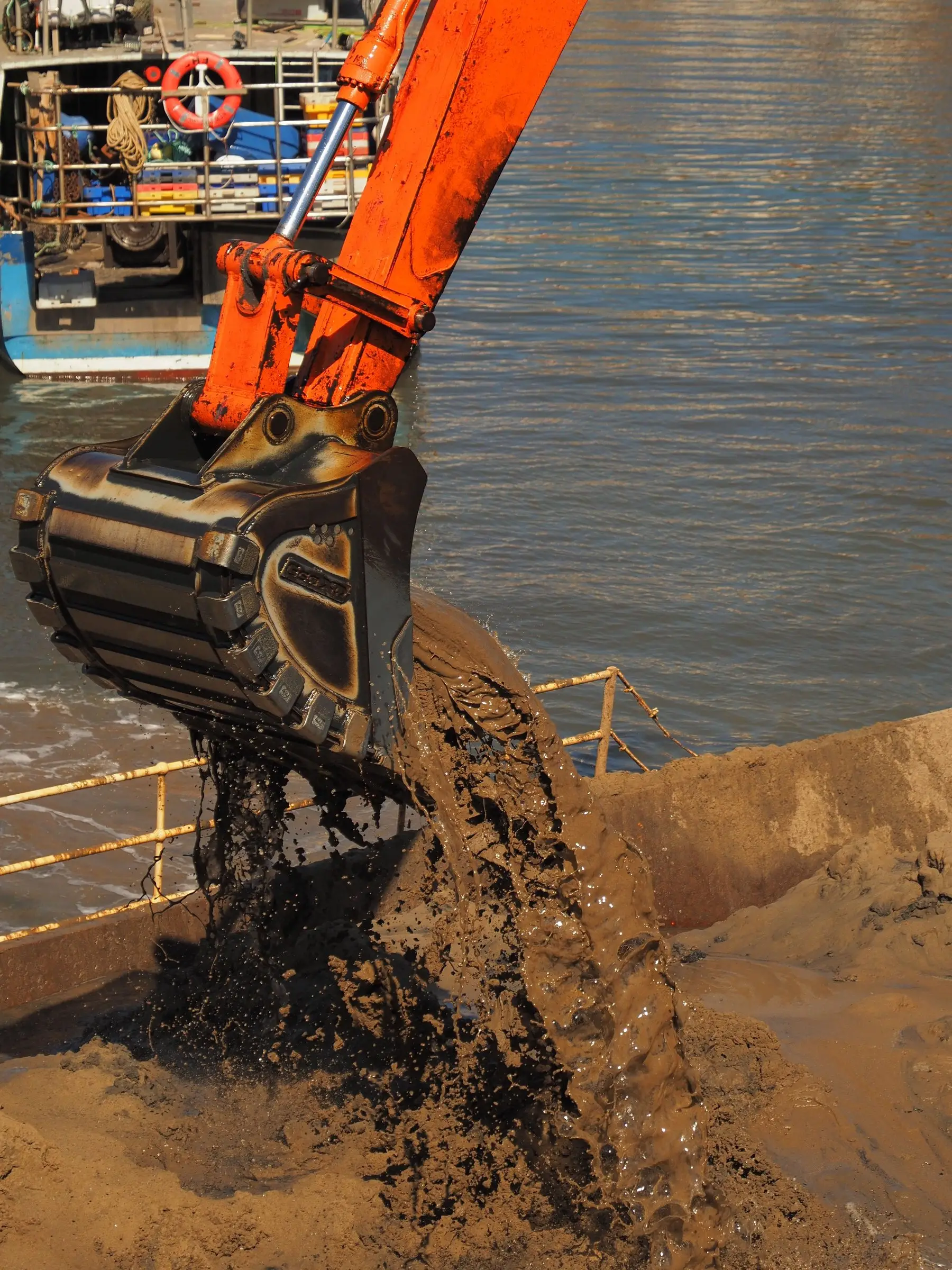 An excavator bucket tipping wet grey silt onto a bank during harbour dredging