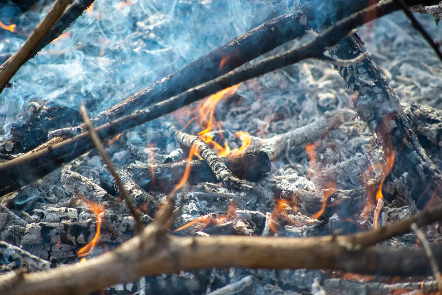 A fire burned down to a bed of grey ash and charred wood with low flames and smoke still rising