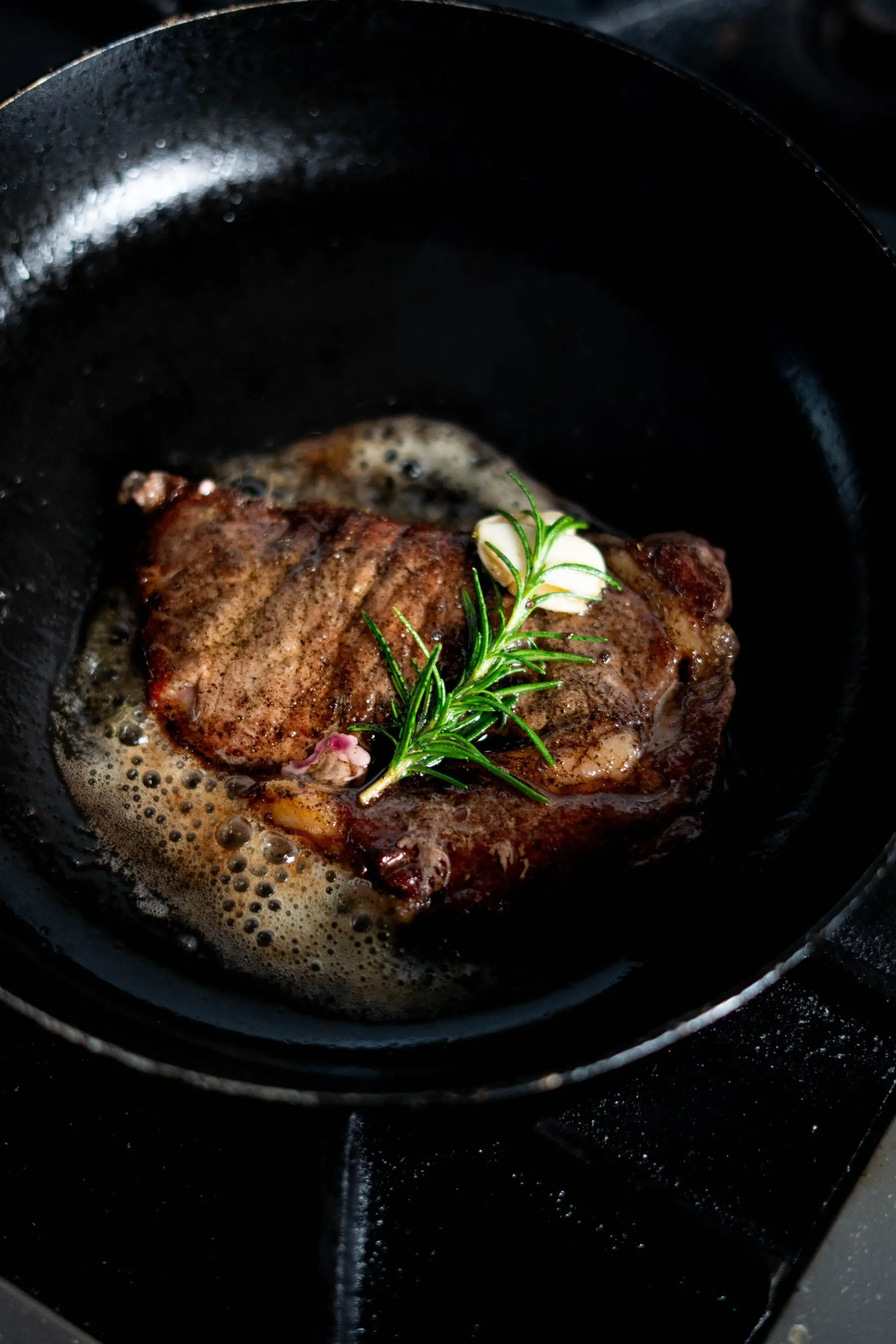 A steak searing in a smooth cast-iron skillet with butter, rosemary and garlic, crust forming on the underside