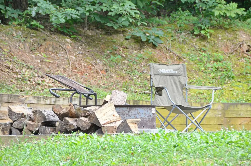 Split firewood piled beside a steel campground fire ring with a cooking grate, camp chair alongside