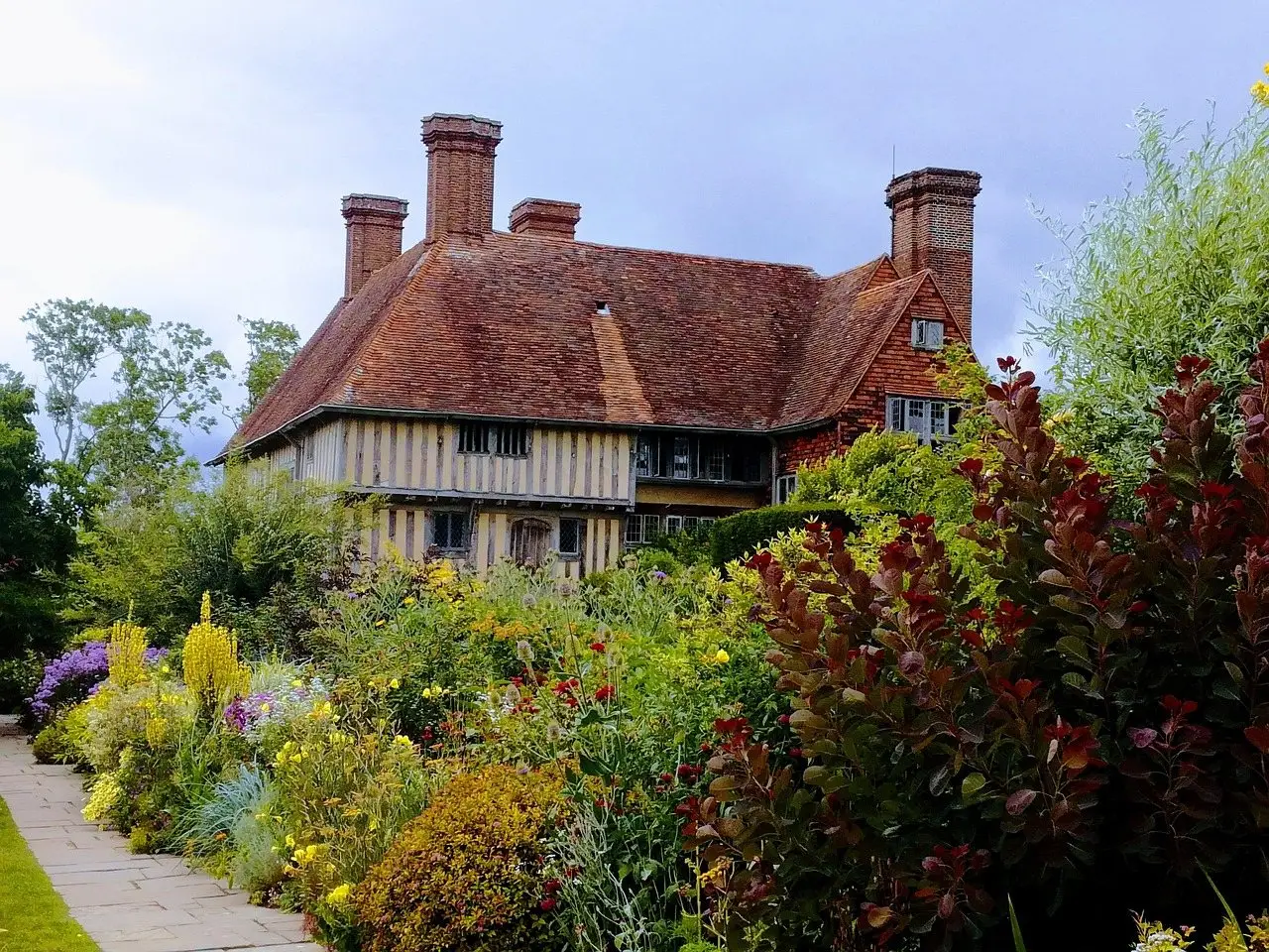 A deep mixed flower border of perennials and shrubs running along a path in front of a timbered house