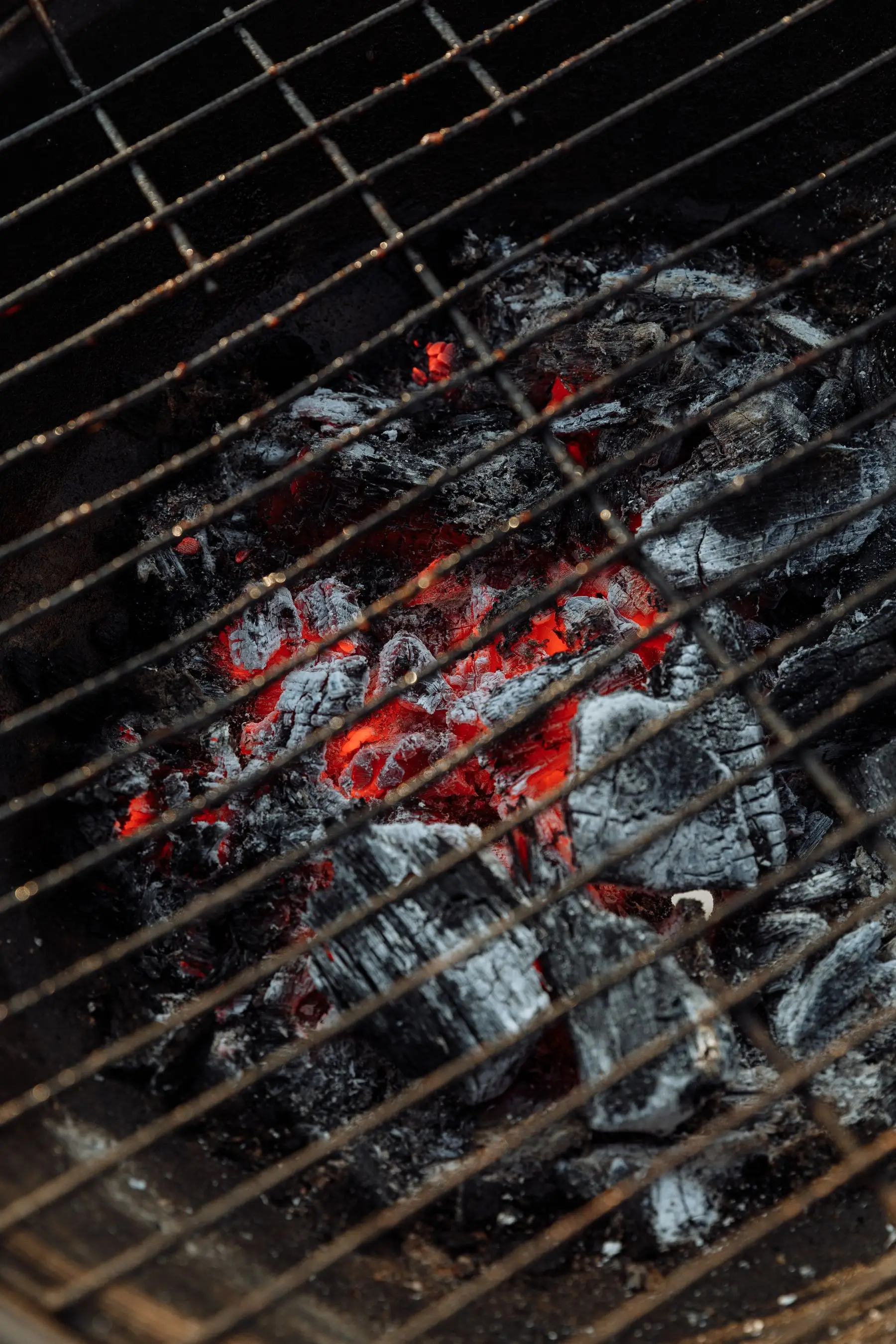 Charcoal under a grill grate, mostly covered in grey ash with red embers still glowing beneath it