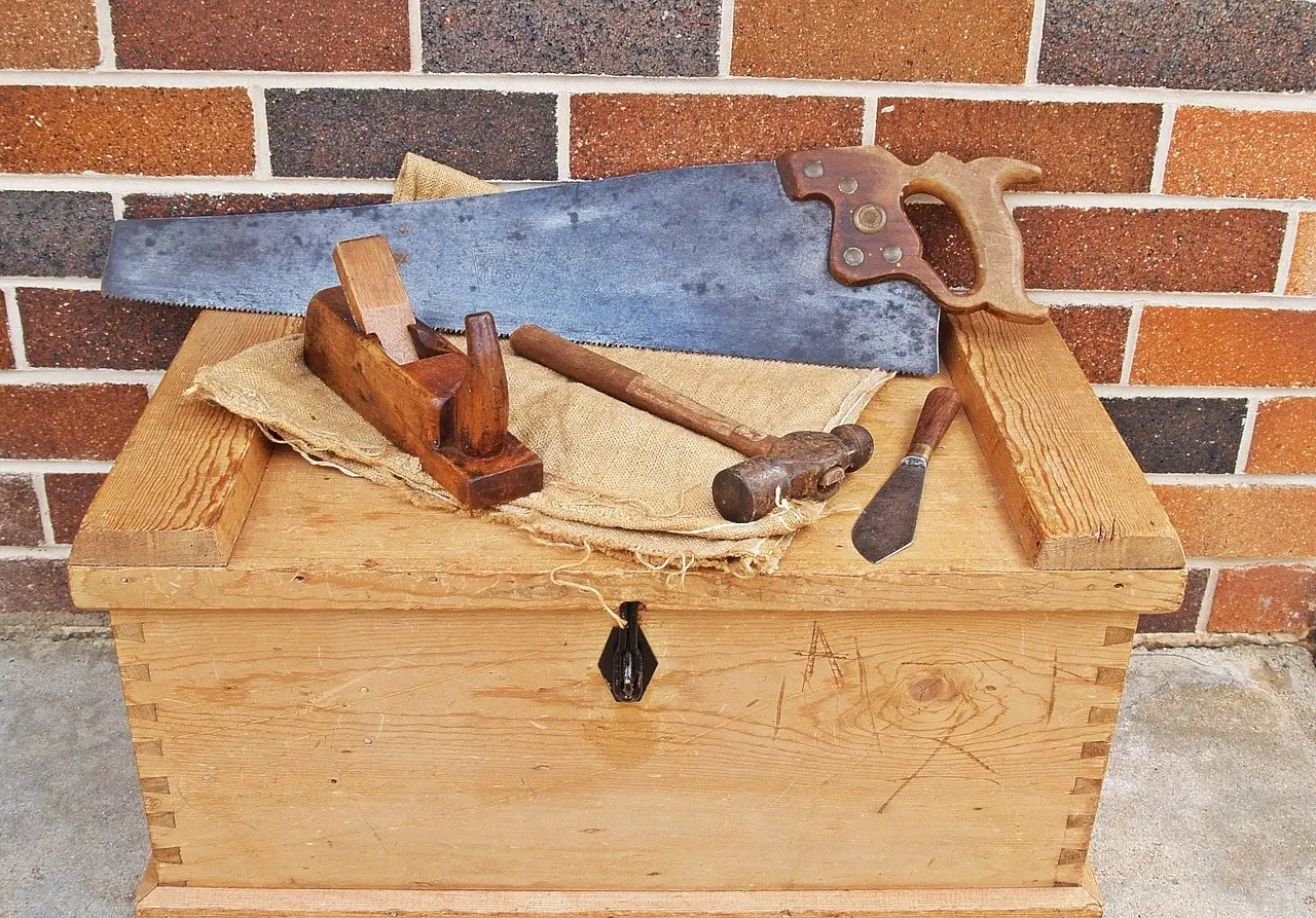 A wooden carpenter's tool chest with a hand saw, plane, mallet and chisel laid across the lid
