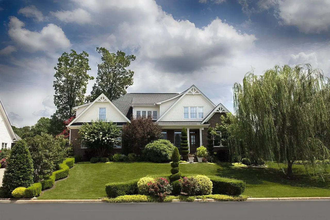 A suburban house behind a mown lawn with clipped shrubs and a willow tree