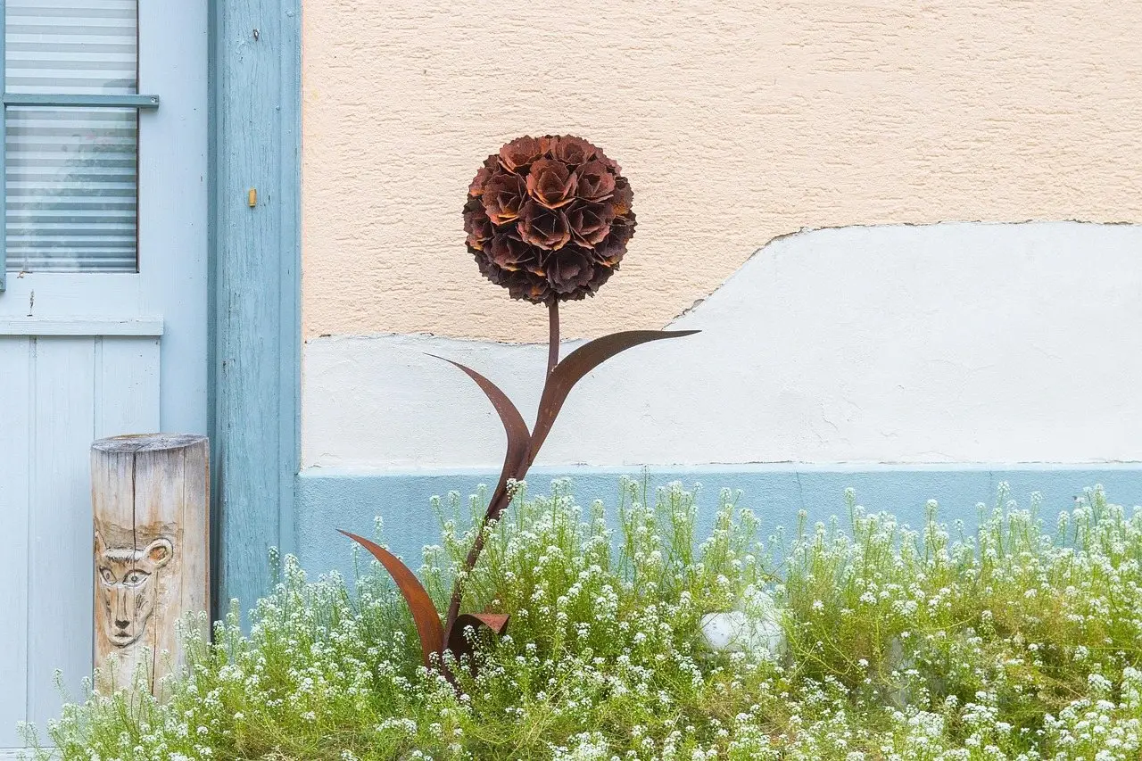 A rusted steel flower sculpture standing in planting against a pale rendered wall