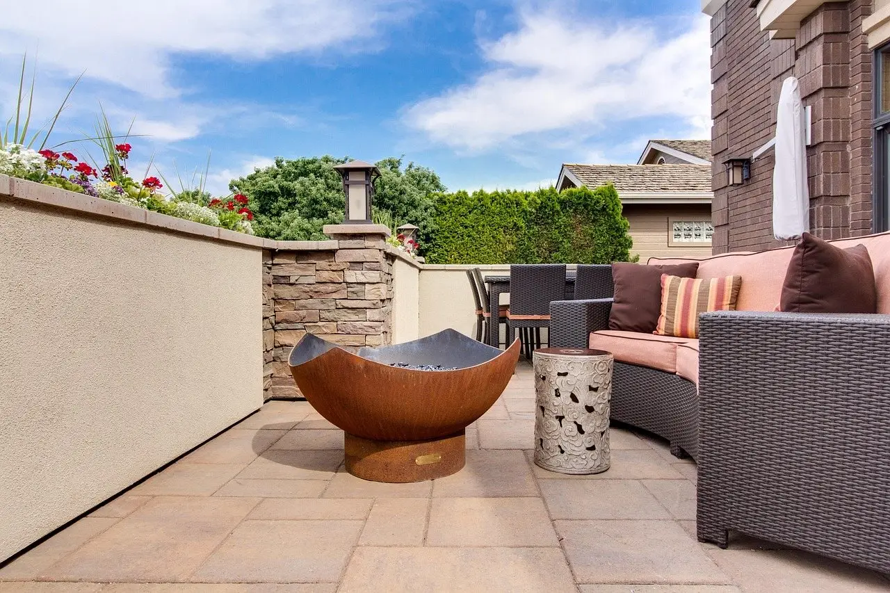 A paved patio with a corner sofa and side table arranged around a copper fire bowl