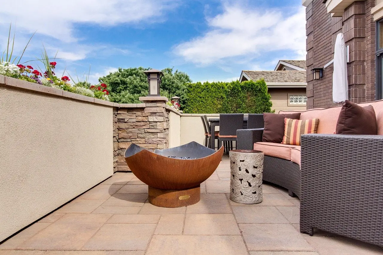 A paved patio with a corner sofa and side table arranged around a copper fire bowl