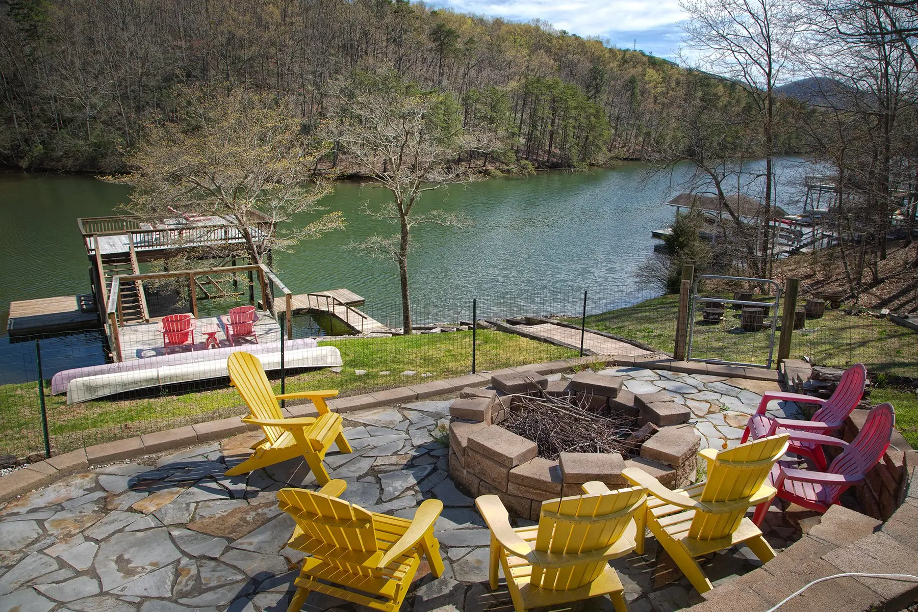A stone fire pit ringed by Adirondack chairs on a lakeside patio
