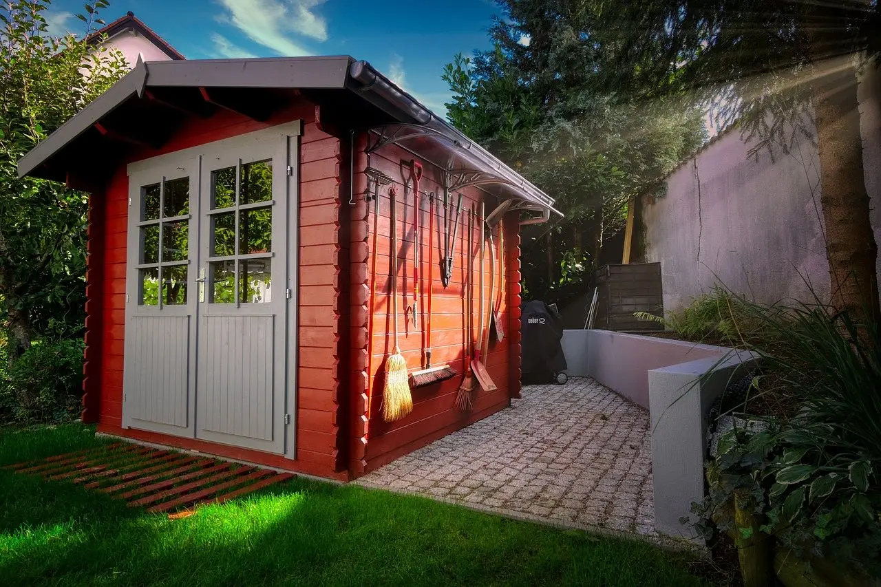 A red timber garden shed with tools hung on its side wall, beside a block-paved path and lawn