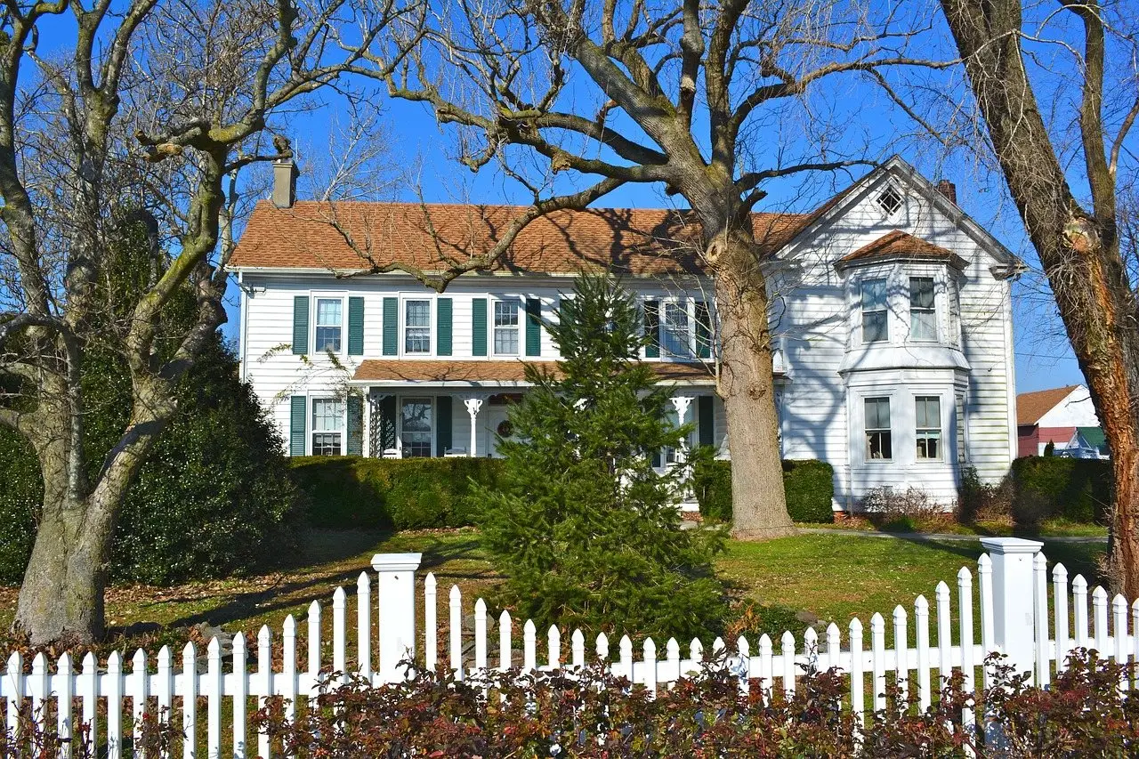 A white clapboard house behind a low white picket fence and mature trees