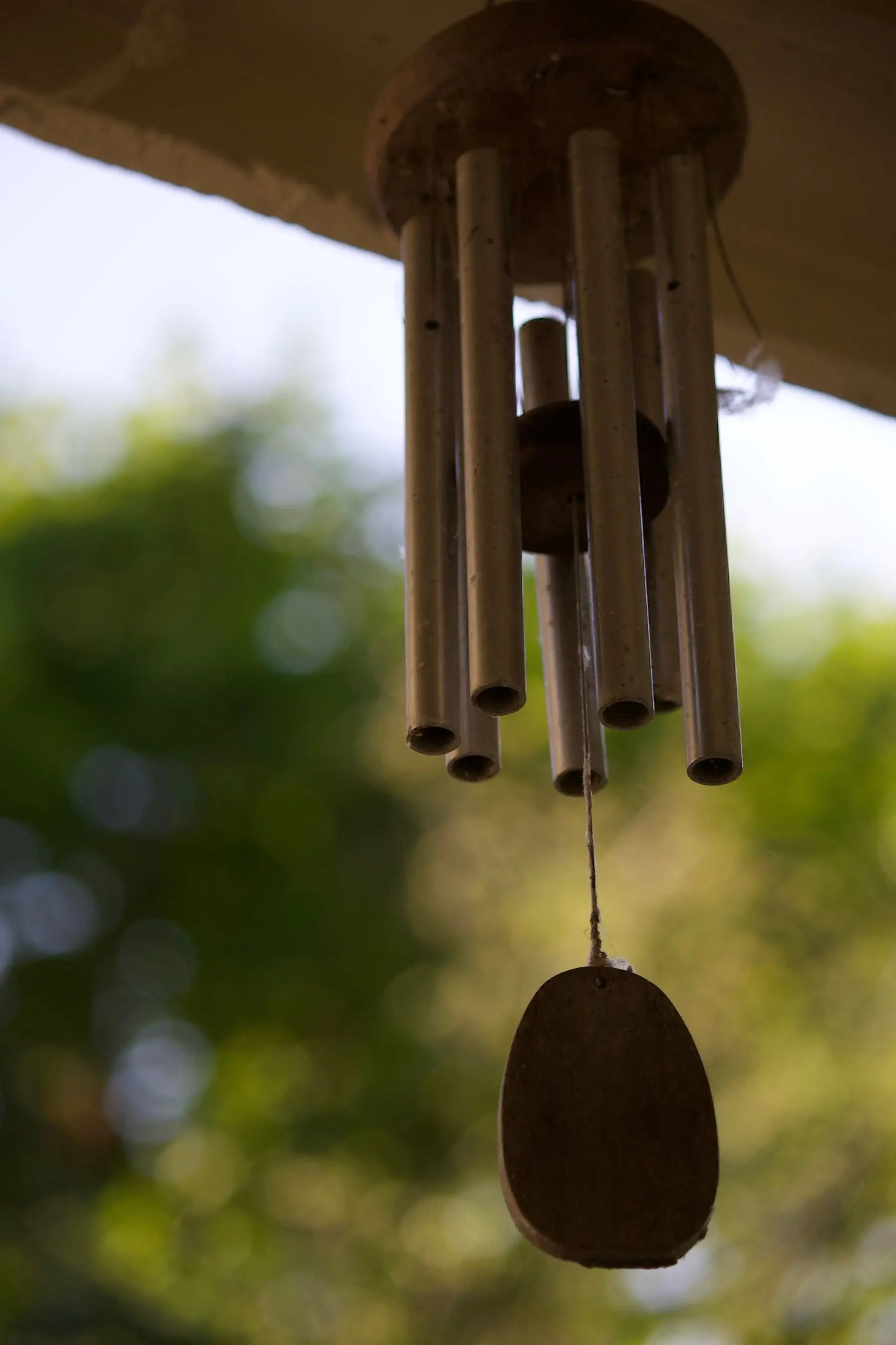 A metal tube wind chime hanging under an eave against sunlit foliage
